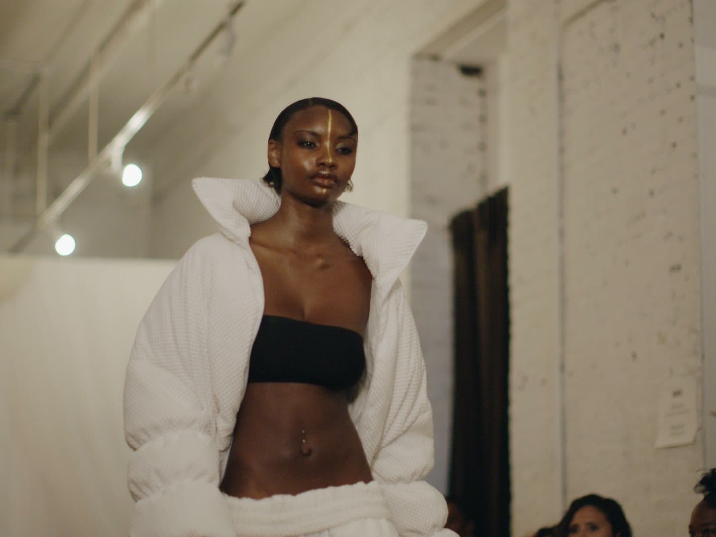 A model in a white high-collared puffer on the Between Worlds runway, a gold line painted down her face